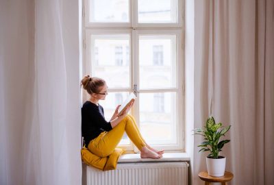 A young female student with a book sitting on window sill, studying.